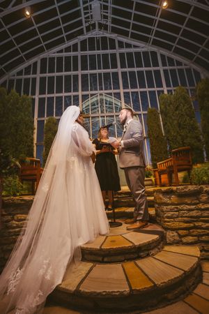 Bride and groom exchange vows at night in a stunning glass conservatory wedding ceremony with lush greenery.