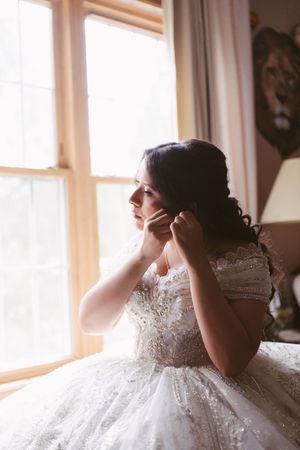 Bride in lace ball gown adjusting earring by sunlit window on wedding day.