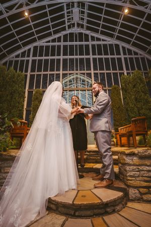 Bride and groom exchanging vows with officiant inside glass greenhouse conservatory at night.