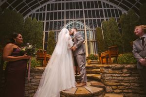 Bride and groom share a first kiss during an elegant indoor greenhouse wedding ceremony with stone steps.