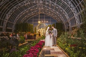 Bride and groom stand in a stunning glass greenhouse wedding venue adorned with flowers and twinkling lights.