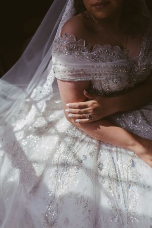 Bride in beaded off-shoulder wedding gown with veil, showcasing diamond ring while seated in soft natural light.