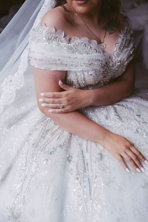 Close-up of a bride in a beaded off-shoulder wedding gown with crossed arms displaying an engagement ring.