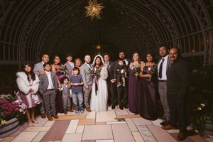 Wedding party group portrait with bride, groom, bridesmaids in purple, and family inside conservatory.