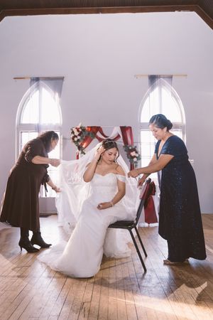 Bride in white gown sits as two women adjust her veil in a bright chapel with arched windows.