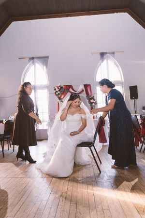 Bride in white gown sits in chair as two women assist with her veil inside a decorated wedding venue.