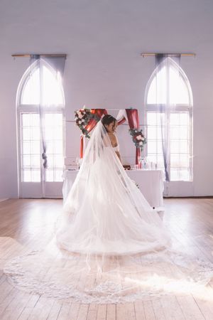 Bride in a flowing white lace wedding gown and long veil standing in a bright white venue with arched windows.