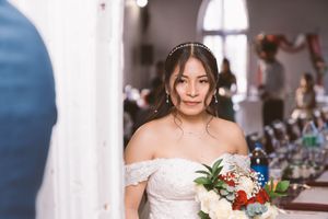 Bride in white off-shoulder lace gown holding floral bouquet with red and white flowers inside a church.