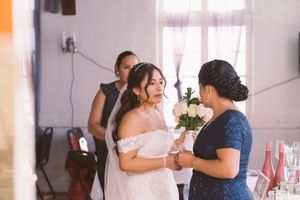 Bride in white off-shoulder gown receiving bouquet of white roses from guest in blue lace dress at wedding reception.