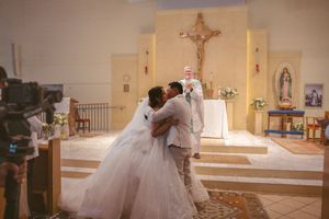 Newlyweds sharing first kiss at church altar with priest officiating Catholic wedding.