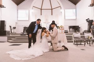 Bride and groom kneeling in prayer with officiant during wedding ceremony inside a white church.