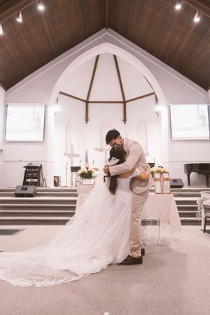 Bride and groom share a romantic dip kiss at the altar of a white church with wooden ceiling.