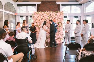 Couple shares first kiss at indoor wedding ceremony with pink floral wall backdrop and wedding party standing alongside.