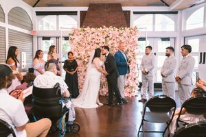 Couple exchanges vows at indoor wedding ceremony before floral wall backdrop surrounded by wedding party.