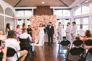 Bride and groom walk down the aisle at indoor wedding ceremony with pink floral backdrop and guests seated in black chairs.