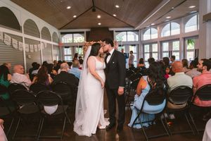 Bride and groom share first kiss at indoor wedding ceremony with guests seated in chairs in elegant venue.