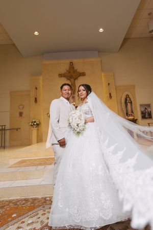 Bride and groom posing in a Catholic church, bride wearing a lace ballgown with flowing veil holding a white bouquet.