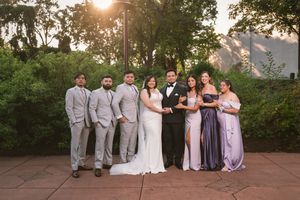 Wedding party poses outdoors at golden hour, with groomsmen in gray suits and bridesmaids in lavender gowns flanking the bride and groom.