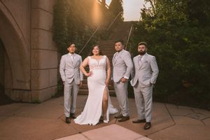 Bride in white gown poses with three groomsmen in gray suits at golden hour outside a stone building.