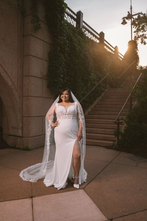 Bride in white lace gown and long veil poses on stone steps at golden hour.