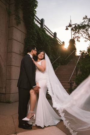 Bride in white mermaid gown and veil embraces groom in black suit at sunset near stone steps outdoors.