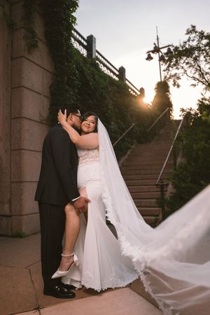Bride and groom share a romantic embrace at sunset near stone steps, her white gown and veil flowing gracefully.