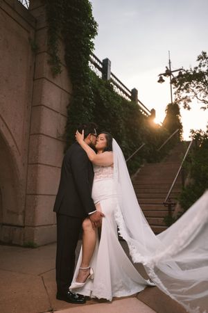 Bride and groom share a romantic kiss at sunset near stone steps with ivy-covered walls, her veil flowing gracefully.