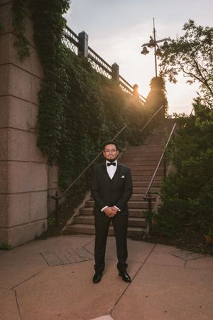 Groom in black tuxedo and bow tie stands confidently before stone steps and ivy-covered walls at sunset.