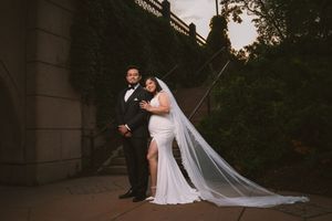 Bride and groom pose elegantly outdoors at dusk against stone wall with cathedral veil trailing.