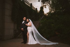 Bride in white gown with long veil and groom in black tuxedo pose together on stone steps at dusk.