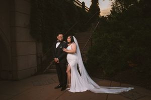 Bride and groom pose elegantly at sunset near stone steps, bride wearing a white gown with long veil and slit.