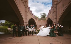 Wedding party poses dramatically under stone bridge arches, with groomsmen lifting the bride in a white gown and green bridesmaid dresses.