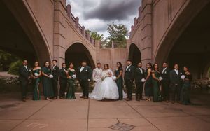 Wedding party in green and black attire posing under stone arches with bride and groom centered.