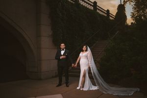 Bride in white gown with long veil and groom in tuxedo hold hands on stone steps at sunset wedding.