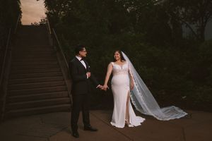 Bride and groom hold hands near stone staircase at dusk, bride wearing fitted gown with long veil.