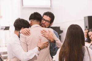 Man with glasses shares a warm group hug with guests at a wedding celebration indoors.