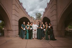 Groom in white suit with bridesmaids in emerald green gowns posing under stone arches at outdoor wedding venue.
