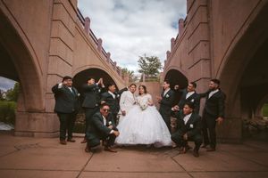 Wedding party poses under stone bridge arches, bride in white gown with groom and groomsmen in dark suits.