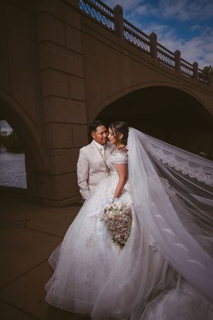 Bride and groom posing under stone bridge with flowing lace veil and bouquet.