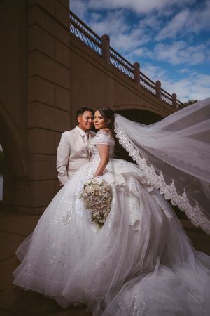 Bride and groom embracing under stone bridge with dramatic flowing lace cathedral veil.