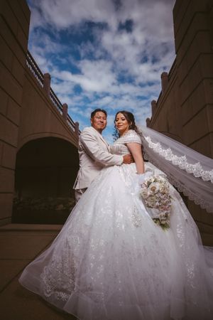 Bride in white lace ball gown with flowing veil and groom in tan suit pose on elegant staircase under blue sky.