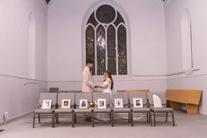 Couple exchanging vows in a small chapel with stained glass window, framed photos on chairs honoring absent loved ones.