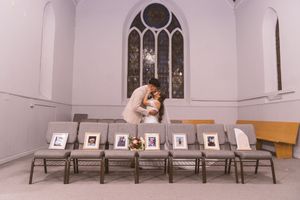 Bride and groom share a romantic kiss at the altar of a church chapel with framed photos displayed on chairs.