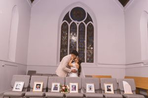 Couple sharing a romantic kiss at the altar of a white chapel with stained glass window and framed photos on chairs.
