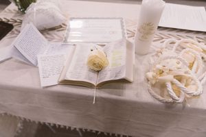 Wedding ceremony table with open vow book, floral boutonniere, unity candle, and decorative rope on white linen.