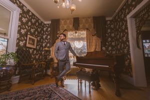 A well-dressed man in a gray suit stands beside a grand piano in an elegant vintage room with ornate wallpaper and chandelier.