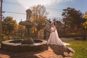 Bride and groom posing by ornate stone fountain in autumn garden setting on their wedding day.