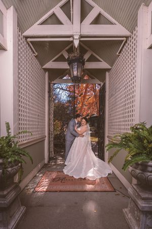 Bride and groom embrace under elegant white pergola entrance with autumn foliage backdrop at romantic outdoor wedding venue.
