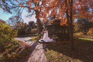 Bride and groom embrace under vibrant autumn foliage near a gazebo in a scenic park on their wedding day.