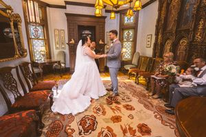 Bride and groom exchange vows in an elegant Victorian-style room with ornate decor and seated guests.